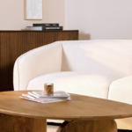 Een gezellige hoek in de woonkamer met een moderne witte bank, een houten salontafel met een paar opgestapelde boeken en een klein kaarsje. Op de achtergrond staat een houten dressoir met daarop ingelijste kunst en boeken.