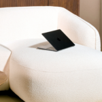 Een strakke zwarte laptop ligt open op het kussen van de Hoekbank Laila - Bouclé Beige rechts, een moderne sectionele bank in een zacht verlichte kamer met houten accenten.