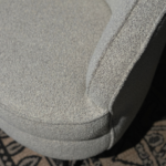 Close-up van een lichtgrijze, getextureerde hoekbank met zachte, afgeronde randen. De bank rust op een patroonkleed, gedeeltelijk zichtbaar in de hoek van de afbeelding, wat contrast toevoegt aan de zachte stof. Subtiele schaduwen versterken de contouren van de bank.