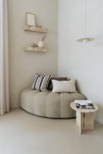Minimalistische kamer met een beige ronde bank, gestreepte en effen kussens, een kleine ronde tafel met een tijdschrift en twee marmeren planken met decoratie-items. Een eenvoudige hanglamp hangt aan het plafond naast doorschijnende gordijnen.