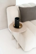 Een minimalistische woonkamerhoek met een beige bank met een donkergrijs kussen. Ernaast staat een ronde, lichtgekleurde bijzettafel met een zwarte kan op een gouden plaat, tegen een neutrale muur.