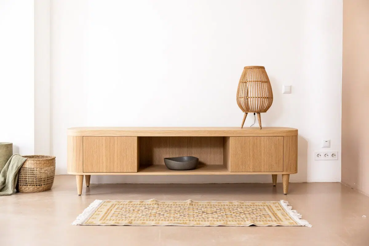 Een minimalistische woonkamerscène met een houten dressoir met een rotanlamp erop. Onder de lamp staat een donkere kom op de middelste plank. Links staat een geweven mand en voor de lichte vloer ligt een patroonkleed.