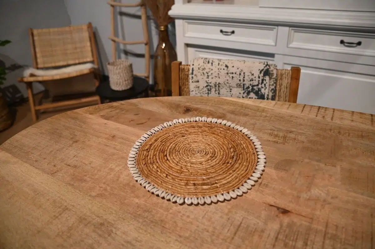 Een ronde houten tafel met een gevlochten rotan placemat in het midden, met een decoratieve schelpenrand. Op de achtergrond staan een houten stoel, een gevlochten stoel, een kleine ronde tafel en een witte kast.