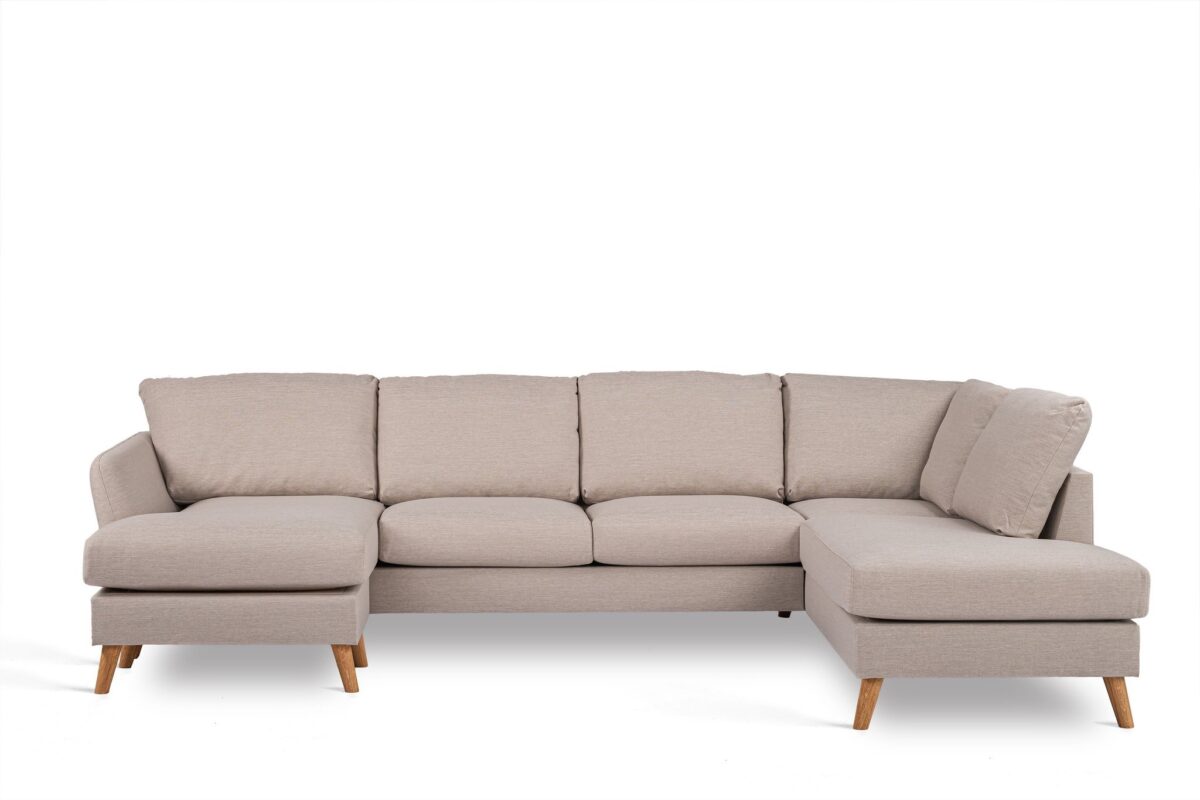 Een lichtbeige L-vormige sectionele bank met houten poten. De bank heeft vijf gestoffeerde zitplaatsen en een chaise longue aan de linkerkant, tegen een effen witte achtergrond.