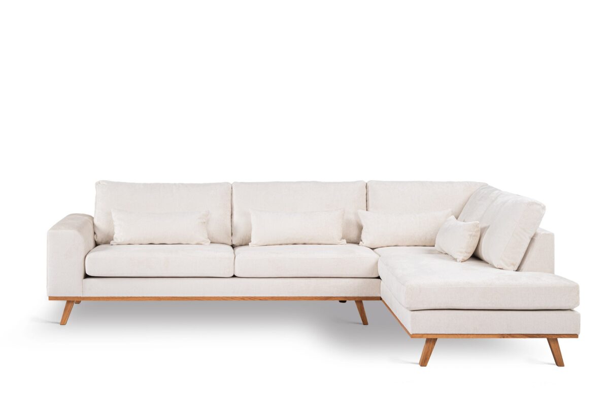 Beige L-vormige hoekbank met houten poten, drie zitkussens, diverse rechthoekige rugkussens en een chaise longue aan de rechterkant, tegen een witte achtergrond.