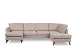 Een beige L-vormige sectionele bank met zwarte poten. De bank heeft vijf gestoffeerde zittingen en een chaise longue aan de linkerkant. Het ontwerp is modern en geschikt voor een woonkamer.
