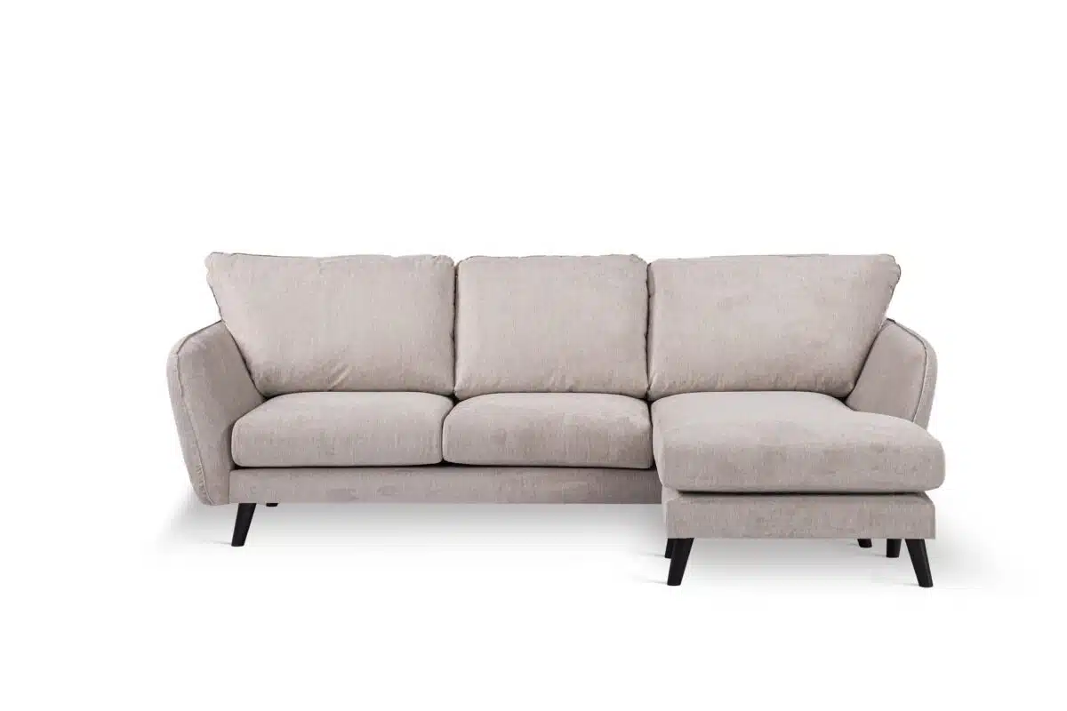 Een lichtbeige sectionele bank met drie kussens en een L-vormige chaise longue aan de rechterkant. Hij heeft zwarte poten en staat op een effen witte achtergrond.