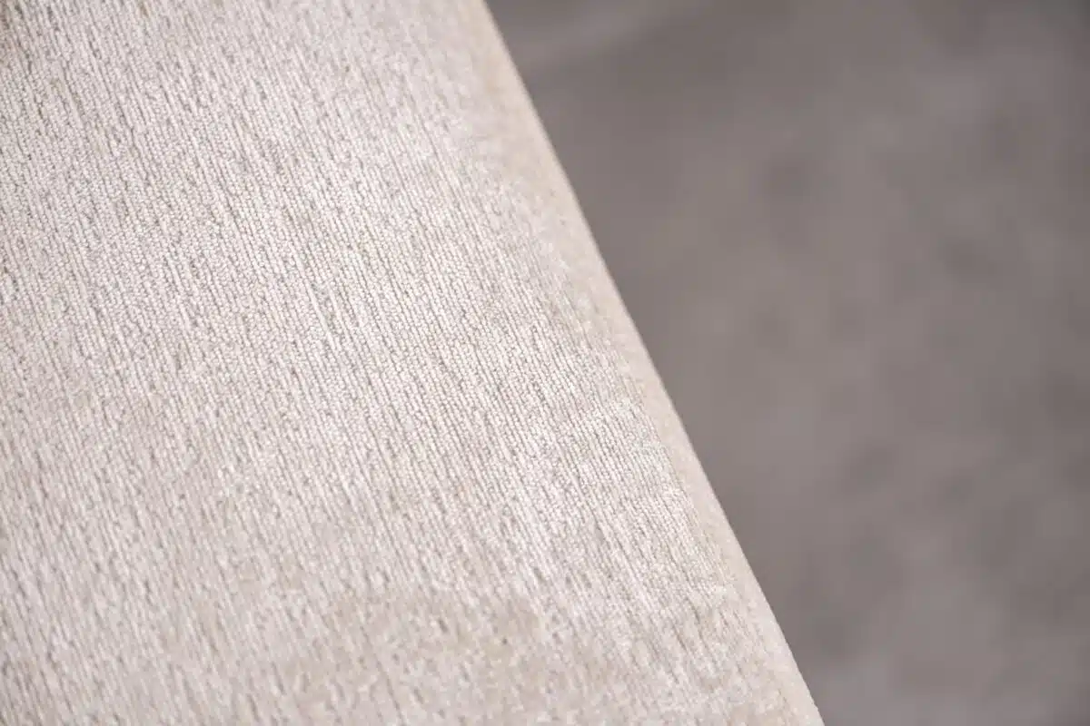 Close-up van een beige stof met een subtiele, gestructureerde weving. Het materiaal heeft een zachte uitstraling en het licht benadrukt het delicate patroon. De achtergrond is onscherp, waardoor de textuur en kleur van de stof worden benadrukt.