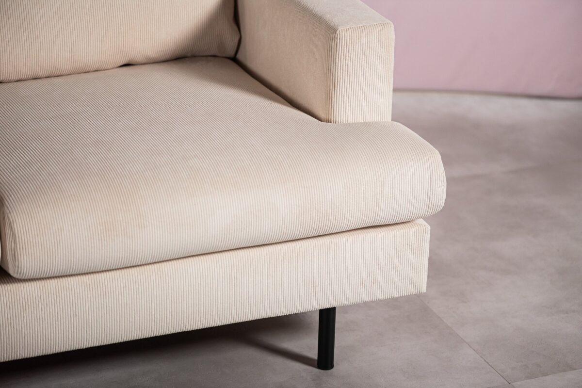 Close-up van een beige corduroy bank in een minimalistische kamer. De bank heeft een licht verzonken armleuning en staat op dunne zwarte poten. De vloer is van lichte stenen tegels en de achtergrond is een zachtroze muur.