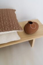 Twee kussens met textuur, een bruine en een beige, zijn op een lichte houten bank gestapeld. Ernaast staat een bruine keramische vaas met witte lijnpatronen, wat een eenvoudig en elegant decorarrangement creëert.