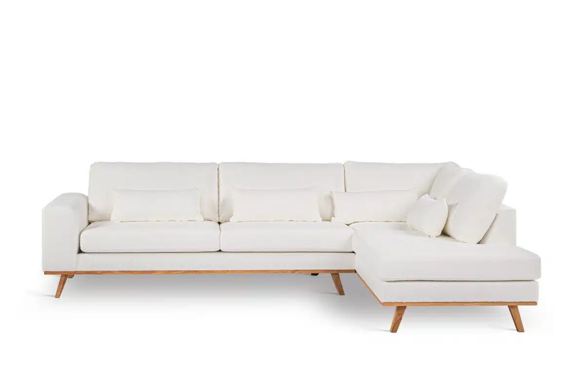 Een moderne witte sectionele bank met een houten basis en poten, met drie zitkussens, een chaise longue aan de rechterkant en vijf bijpassende witte rugkussens. De achtergrond is effen wit.