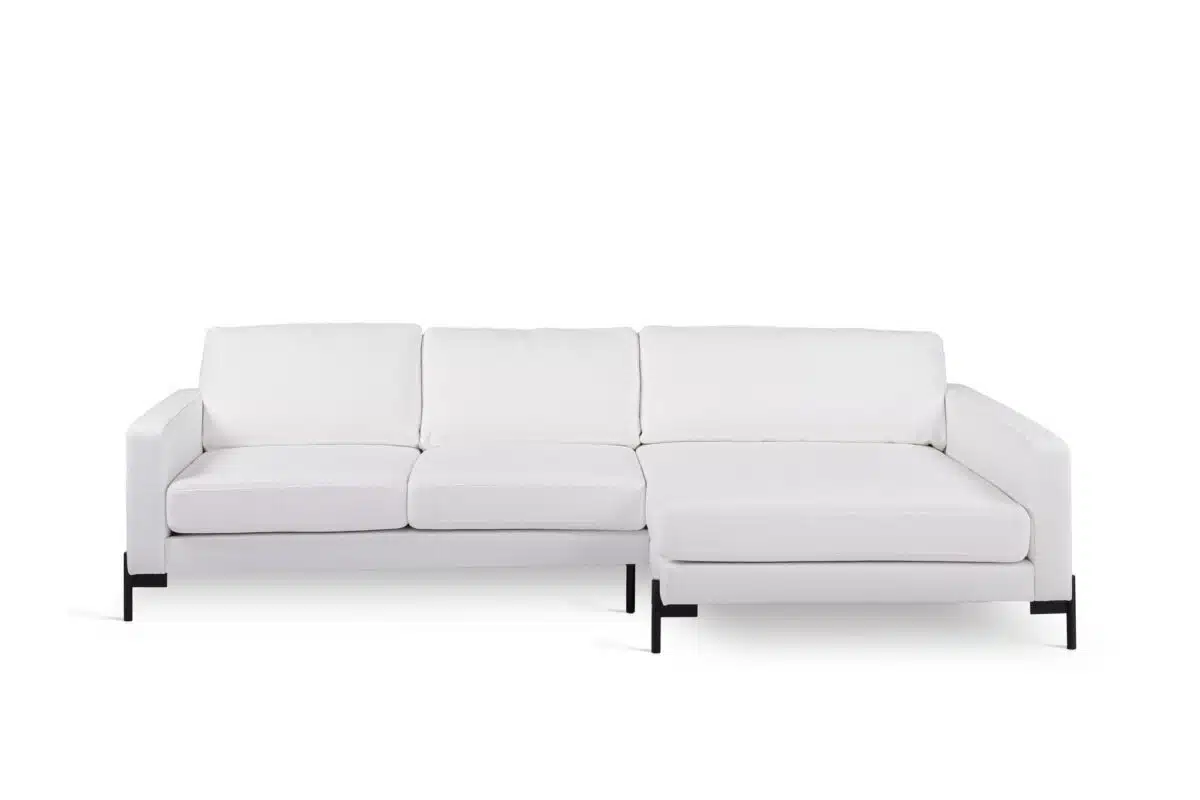 Een moderne witte sectionele bank met drie kussens en een chaise longue aan de rechterkant. Het heeft een strak design met armleuningen en wordt ondersteund door zwarte metalen poten, tegen een effen witte achtergrond.