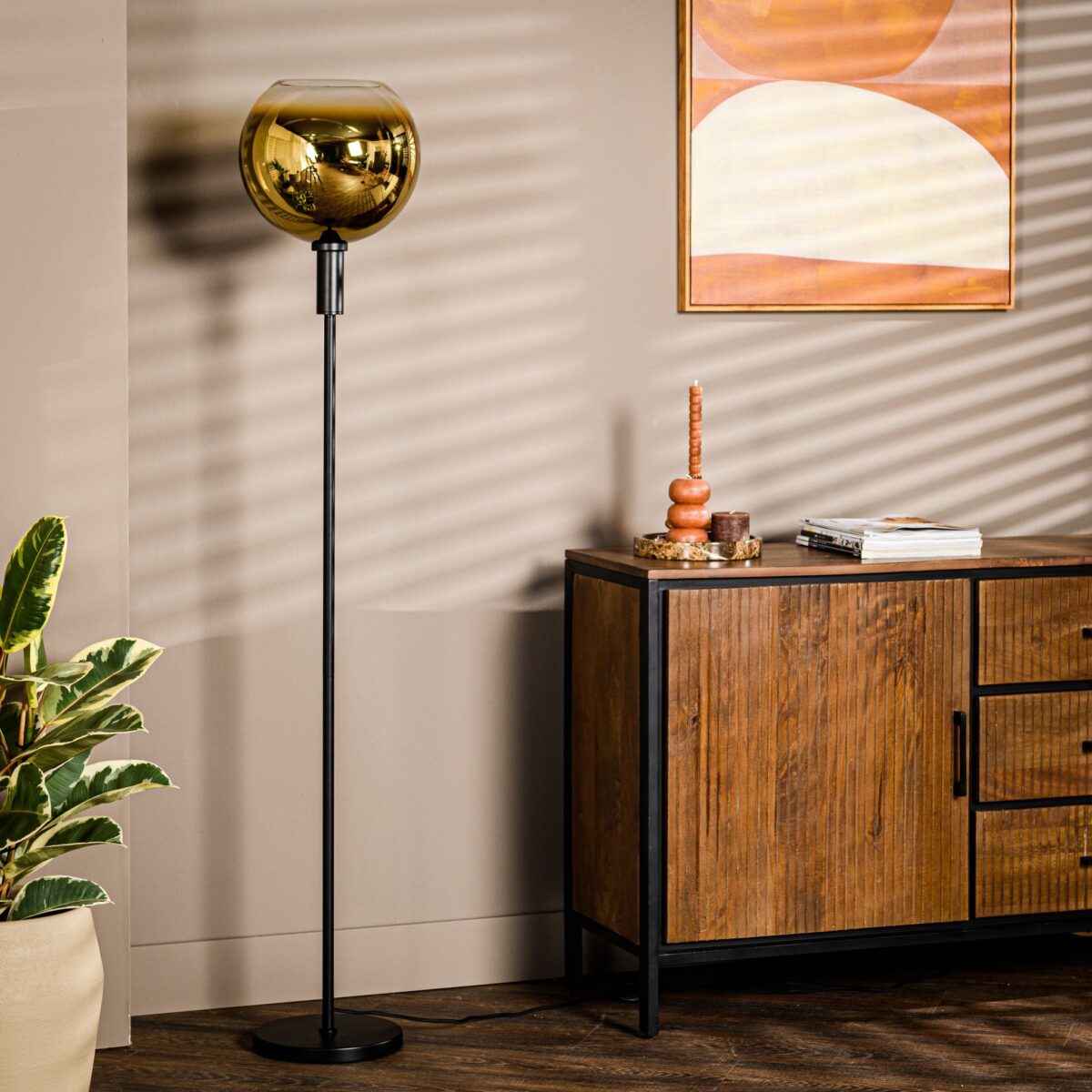 Een kamer met een houten kast, een hoge zwarte staande lamp met een gouden glazen kap en een plant. Aan de muur hangt een ingelijst abstract kunstwerk met gebogen vormen. De kamer heeft een warme, moderne esthetiek.