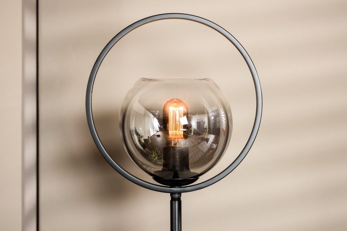 Een moderne lamp met een zichtbare gloeilamp is omsloten door een transparante glazen bol, die wordt omlijst door een cirkelvormige metalen structuur. De lamp is geplaatst tegen een neutrale muur met zachte gestreepte schaduwen.