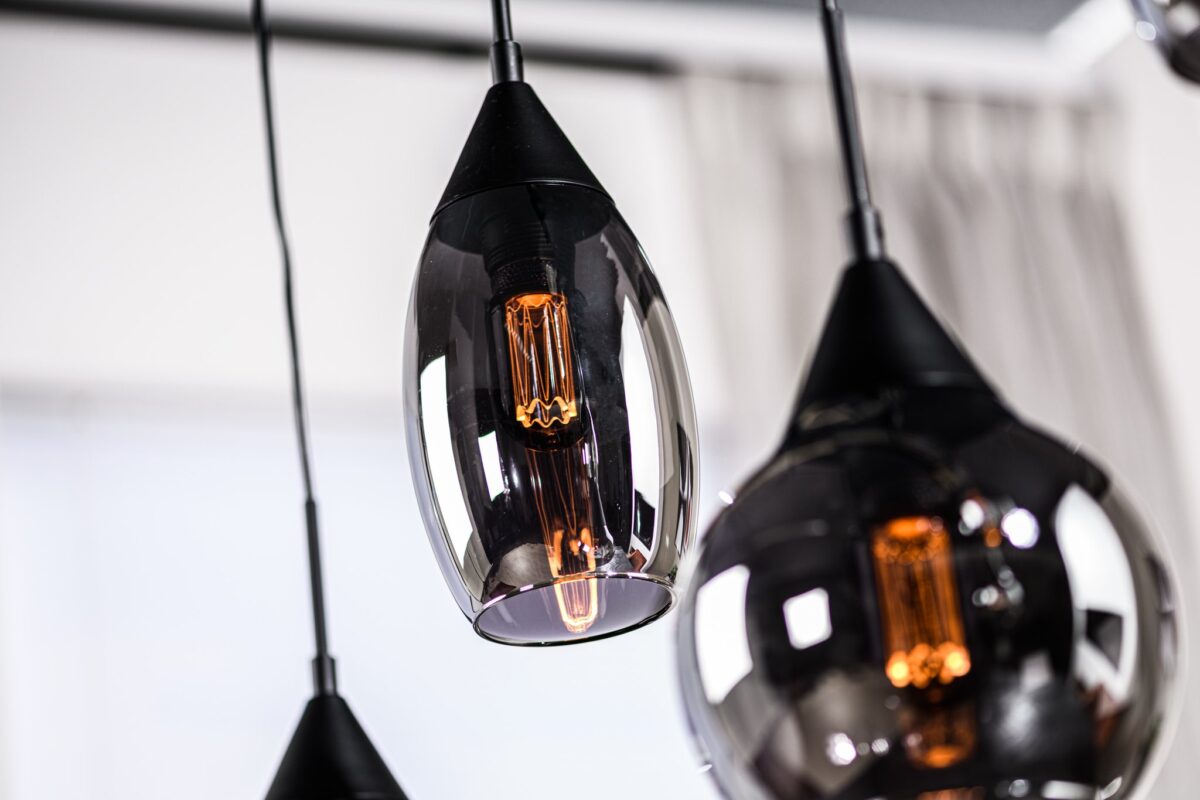 Close-up van drie moderne hanglampen met zwarte glanzende kappen en warm gloeiende filamentlampen. De lampen hangen op verschillende lengtes, met een zachte focusachtergrond van een wit gordijn.