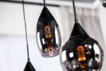 Close-up van drie moderne hanglampen met zwarte glanzende kappen en warm gloeiende filamentlampen. De lampen hangen op verschillende lengtes, met een zachte focusachtergrond van een wit gordijn.