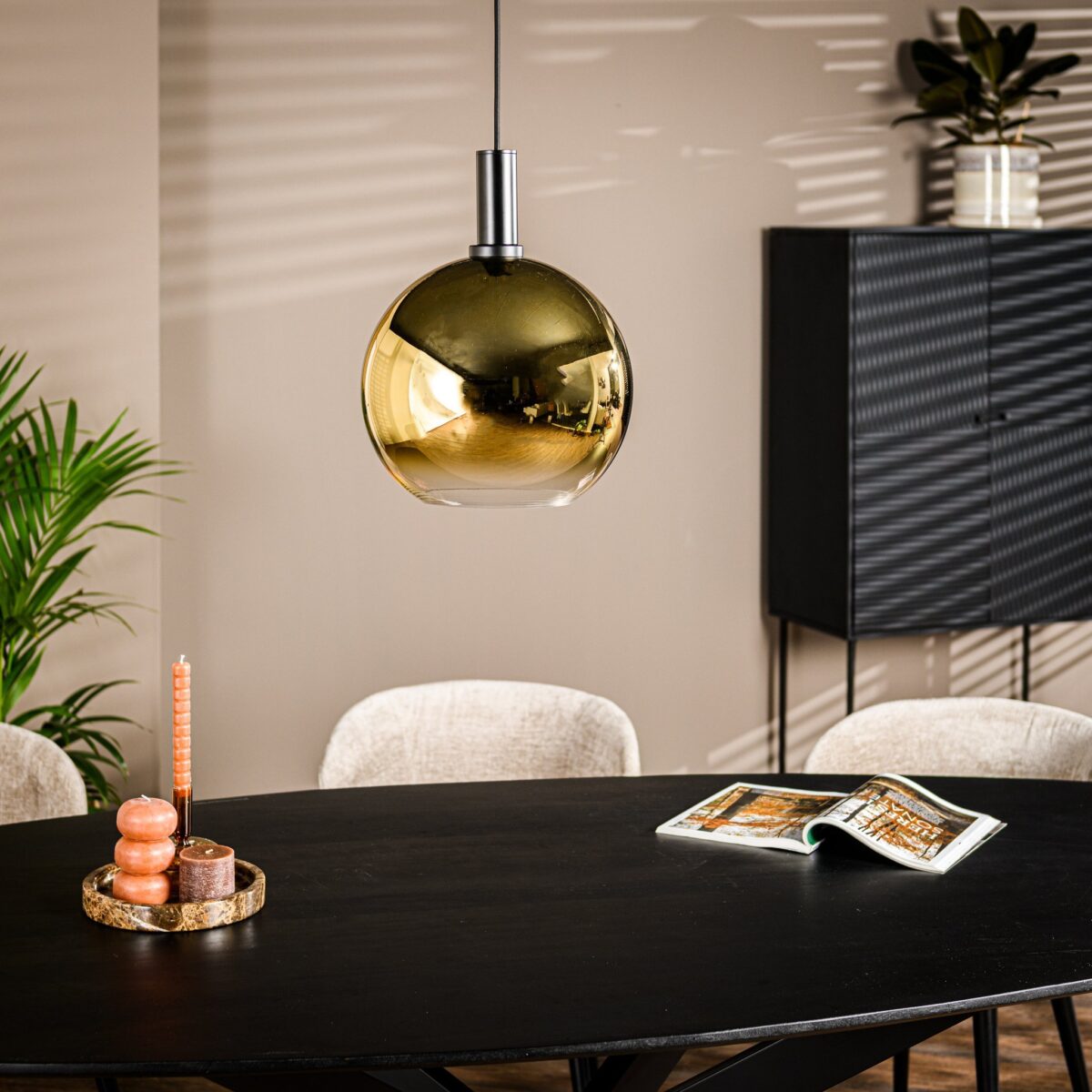 Een moderne eetkamer met een zwarte ovale tafel, beige stoelen en een gouden bolvormige hanglamp. Op de tafel staan gestapelde kaarsenhouders en een open tijdschrift. Een zwarte kast en groene planten staan op de achtergrond. Lichtpatronen op de muren.