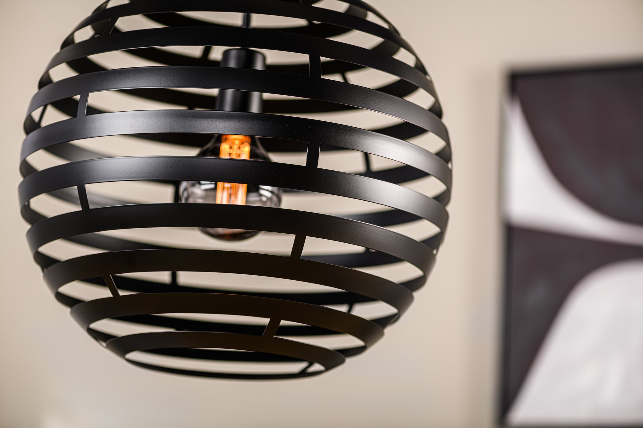 Een close-up van een moderne zwarte bolvormige hanglamp met horizontale lamellen, die een zichtbare lamp binnenin laat zien. De achtergrond is wazig en toont een deel van een zwart-wit abstract kunstwerk op een beige muur.