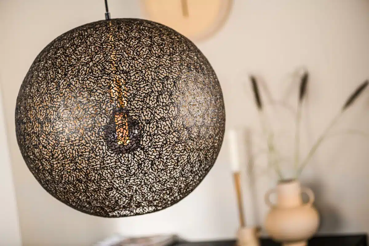 Een close-up van een bolvormige, ingewikkeld gepatroneerde metalen lampenkap met een zichtbare gloeilamp erin. Op de wazige achtergrond staat een beige vaas met groen en een paar kaarsen op een tafel.