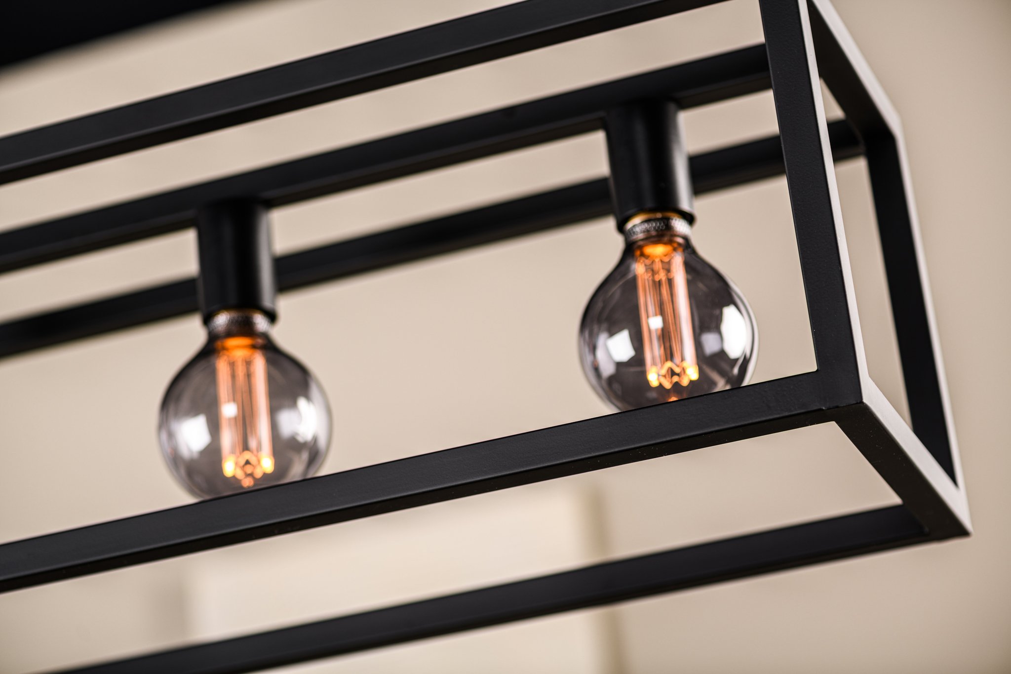 Close-up van twee heldere gloeilampen in een zwart rechthoekig metalen frame, met ingewikkelde filamentdetails. De achtergrond is zacht vervaagd, wat het moderne industriële ontwerp van de lamp benadrukt.