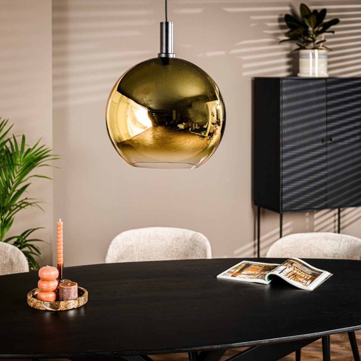 Een moderne eetkamer heeft een zwarte ovale tafel met een dienblad met kaarsen. Boven de tafel hangt een glanzende, gouden bolvormige hanglamp. Op de achtergrond staat een zwarte kast en een plant. Er ligt een open boek op tafel.