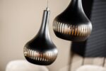 Close-up van twee moderne, zwarte hanglampen met smalle, verticale spleten, die van bovenaf hangen. De achtergrond is onscherp en heeft neutrale tinten die het strakke ontwerp van de lampen benadrukken.