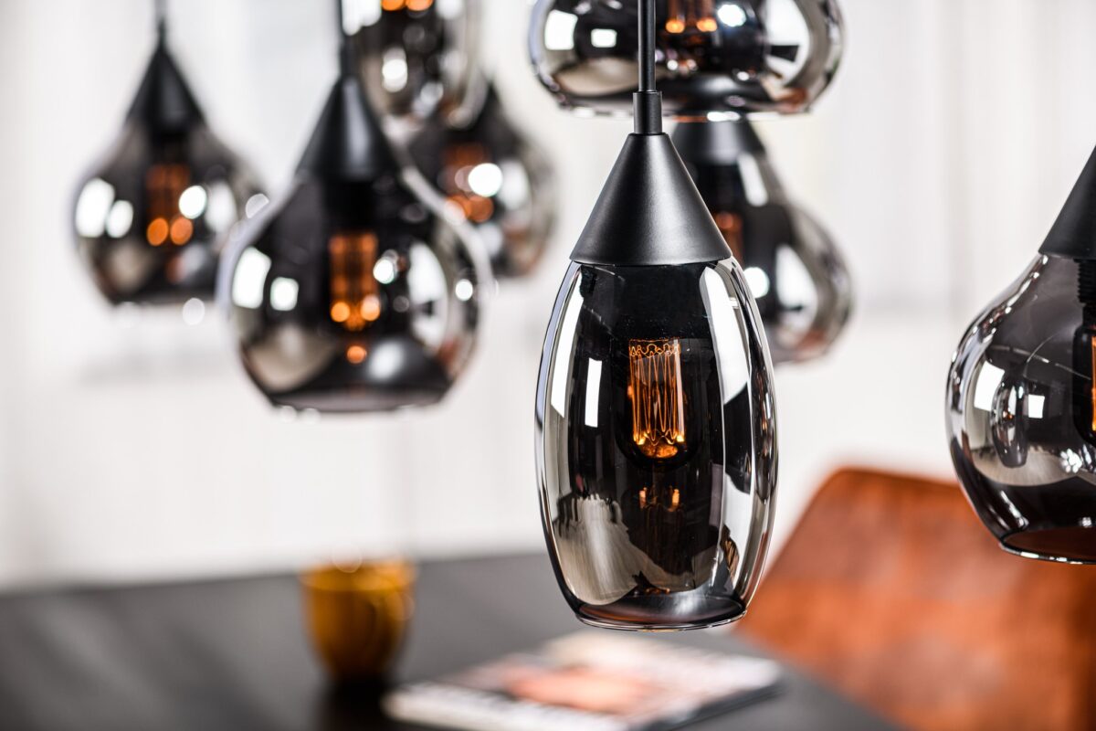 Een cluster van moderne hanglampen met zwarte metalen accenten en heldere glazen kappen hangt boven een tafel. Een oranje stoel en een open tijdschrift zijn zichtbaar op de wazige achtergrond.
