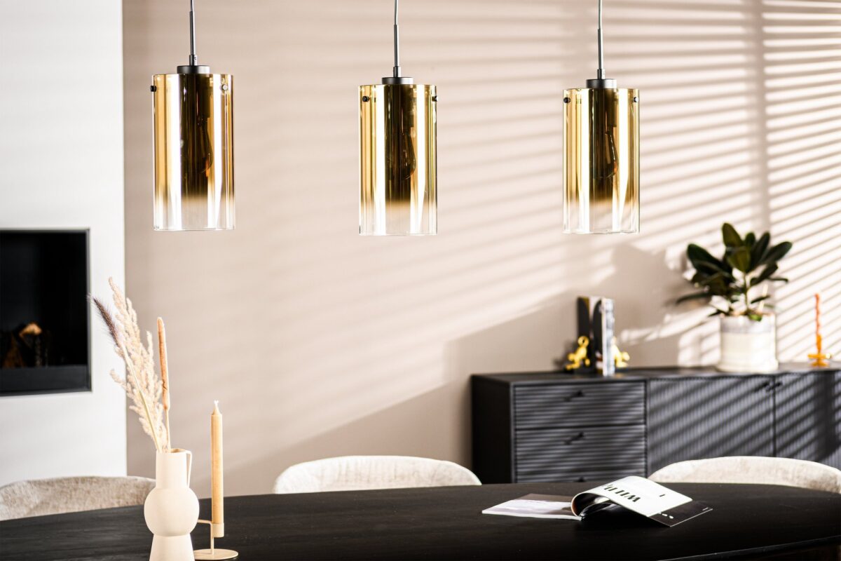 De afbeelding toont een moderne eethoek met een donkere tafel, beige stoelen en een zwart dressoir. Drie gouden cilindervormige hanglampen hangen boven de tafel. Zonlicht creëert gestreepte schaduwen op de witte muur. Een vaas, kaarsen en een tijdschrift staan op de tafel.