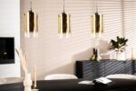 De afbeelding toont een moderne eethoek met een donkere tafel, beige stoelen en een zwart dressoir. Drie gouden cilindervormige hanglampen hangen boven de tafel. Zonlicht creëert gestreepte schaduwen op de witte muur. Een vaas, kaarsen en een tijdschrift staan op de tafel.