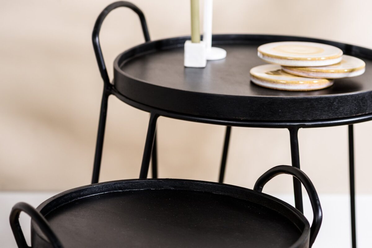 De Salontafel, set van 2, A340 zwart bestaat uit twee ronde zwart metalen bijzettafels met handgrepen. Op de hogere tafel staan een witte kandelaar en ronde onderzetters met goudaccenten tegen een zacht vervaagde neutrale achtergrond.