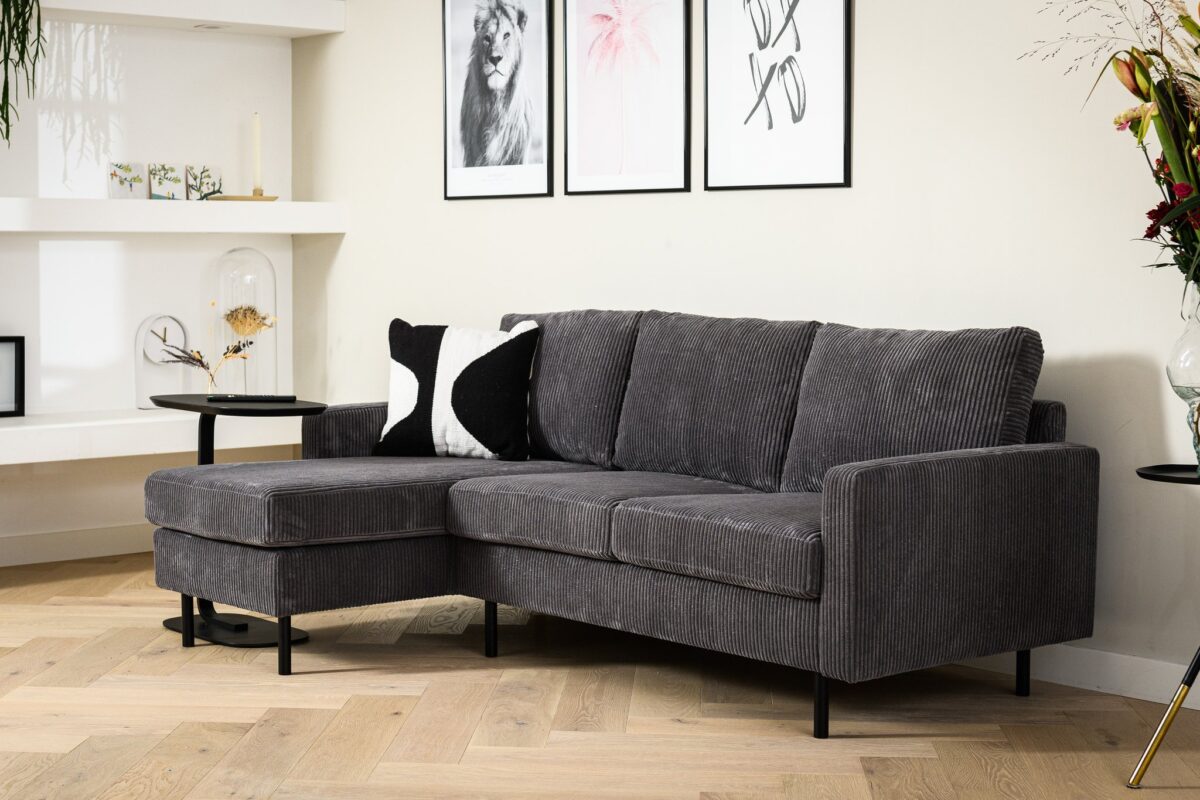 Een moderne woonkamer met de 3 zits bank CL L+R, stof RIB, RIB230 antraciet, gecombineerd met zwarte en witte kussens, een ronde bijzettafel, decoratieve planken, ingelijste muurkunst boven de bank, visgraat houten vloeren en een bloemenvaas.