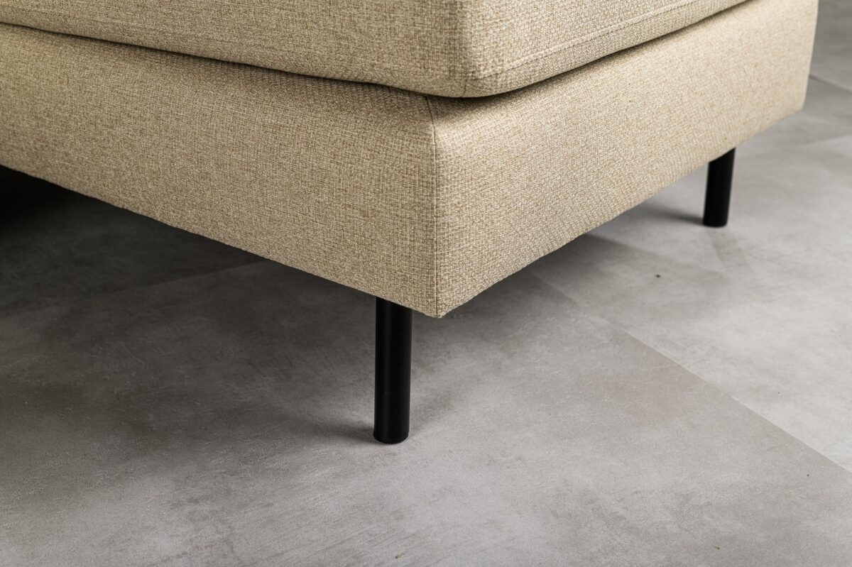 Close-up van de hoek van een beige gestoffeerde bank met zwarte metalen poten op een grijze tegelvloer. De textuur van de stof en het moderne, minimalistische ontwerp zijn zichtbaar.