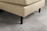 Close-up van de hoek van een beige gestoffeerde bank met zwarte metalen poten op een grijze tegelvloer. De textuur van de stof en het moderne, minimalistische ontwerp zijn zichtbaar.