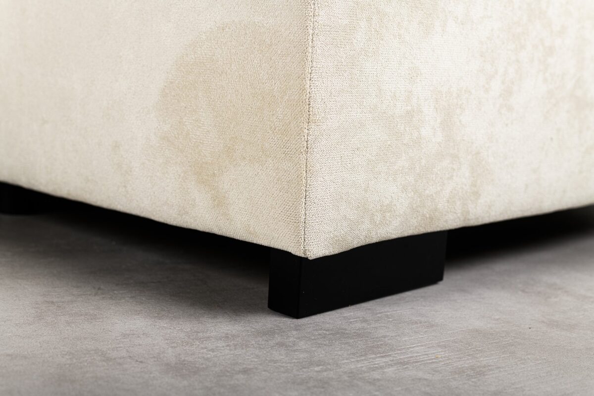 Close-up van de hoek van een beige, gestoffeerde bank met zwarte houten poten op een grijze betonnen vloer.