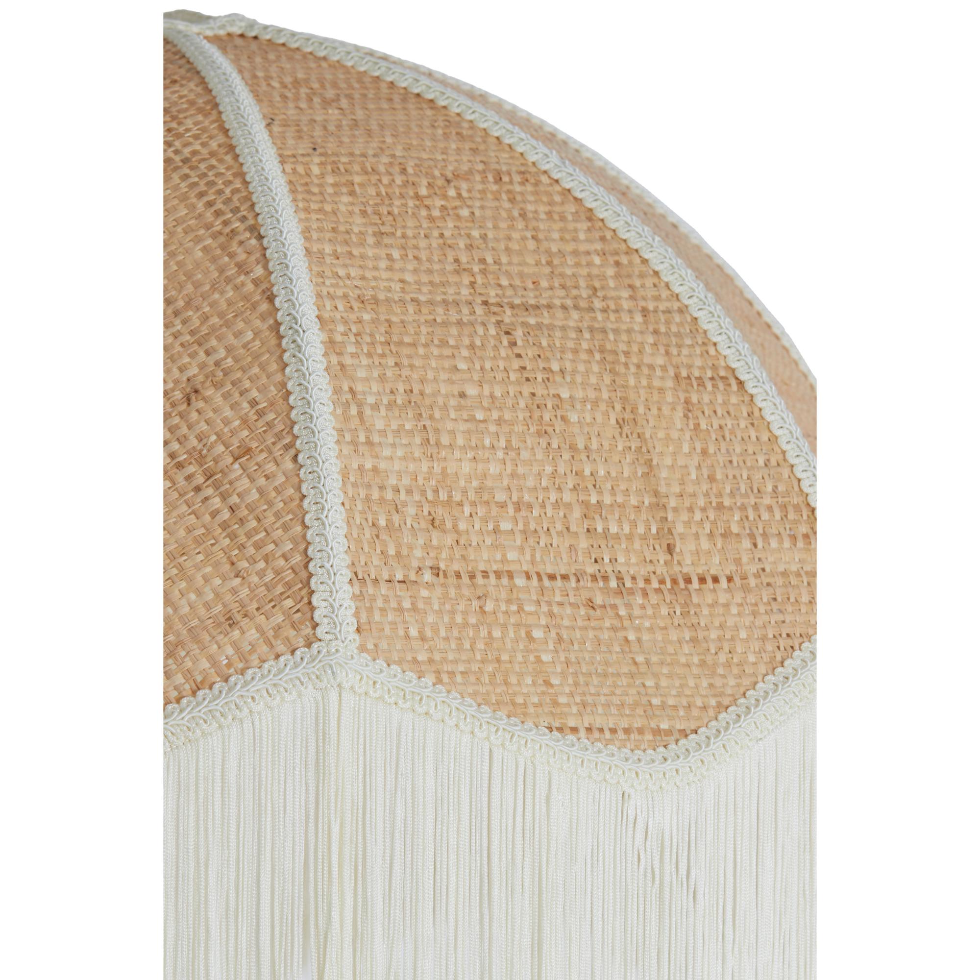 Close-up van een gevlochten rieten lamp met een decoratieve franje langs de rand. De textuur van het riet is gedetailleerd, met verticale en horizontale weefpatronen. De franje voegt een zachte, elegante toets toe aan het rustieke ontwerp.