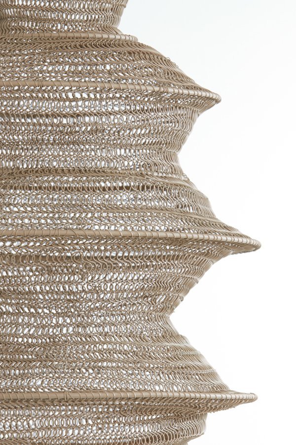 Close-up van een getextureerde, spiraalvormige structuur gemaakt van ingewikkeld geweven, lichtbruin materiaal. Het patroon bevat herhaalde, gelaagde lussen die een golvend, organisch uiterlijk creëren tegen een effen witte achtergrond.