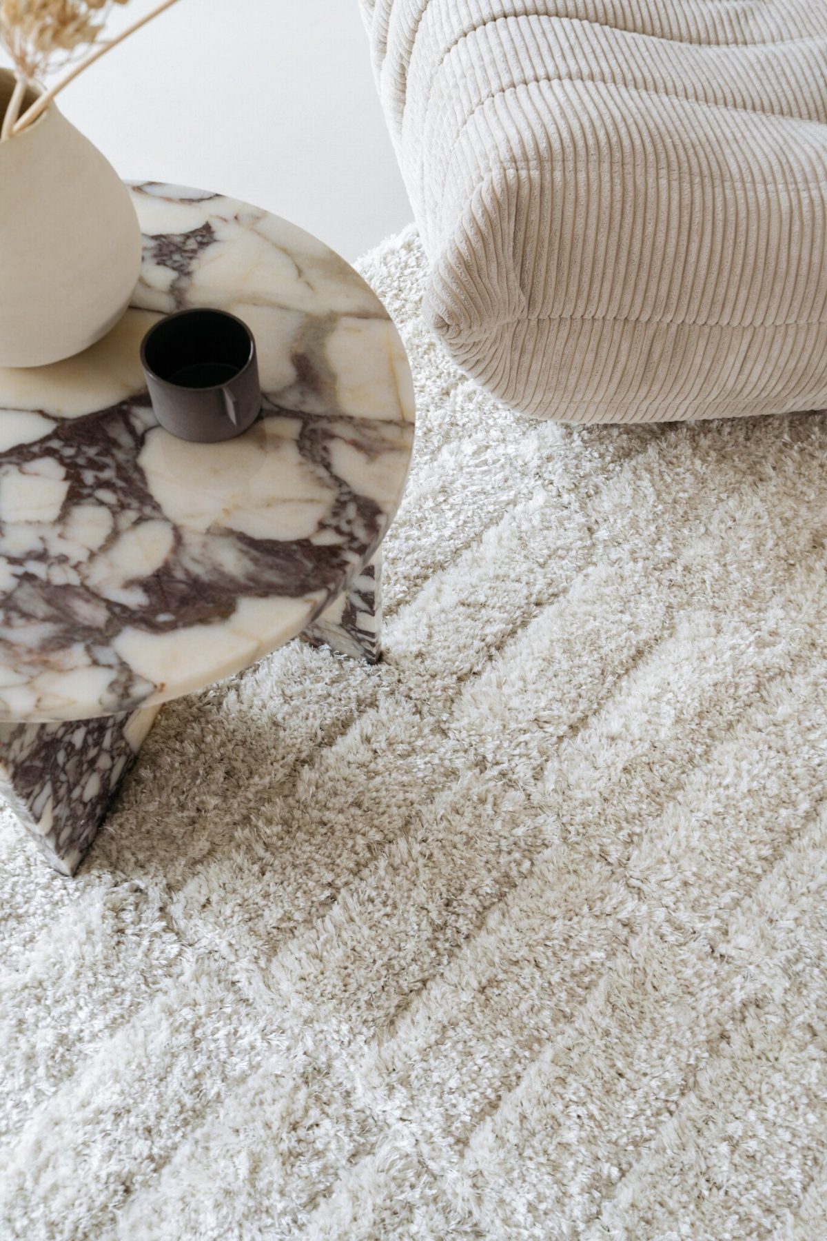 Een gezellige woonkamerhoek met een beige getextureerde bank en een ronde marmeren salontafel met een donkere kop en vaas. Het gebied is versierd met een pluche crèmekleurig tapijt.