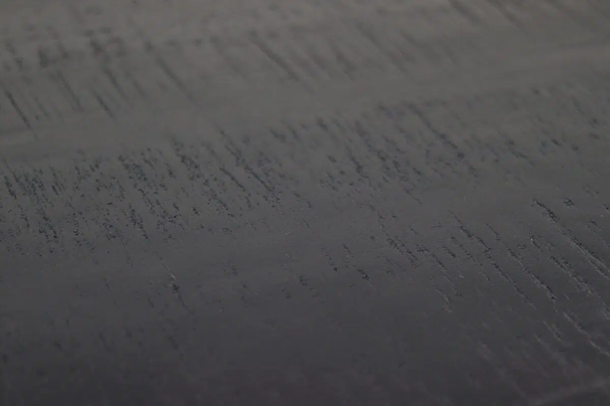 Close-up van een donker getextureerd oppervlak met verticale lijnen en subtiele schaduwvariaties. De textuur lijkt ruw, met onregelmatige patronen die een gevoel van diepte geven.