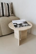 Een kleine ronde houten tafel met een tijdschrift opengeslagen op een zwart-witte pagina. Het staat naast een beige bank met een kussen met zwart-witte strepen. De setting is in een minimalistische kamer met neutrale tinten.