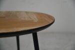 Close-up van een ronde houten tafel met een glad, gepolijst oppervlak en een natuurlijk houtnerfpatroon. De tafel wordt ondersteund door zwarte metalen poten, tegen een effen, lichtgekleurde achtergrond.