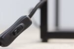 Close-up van een zwart elektrisch snoer met een schakelaar. De achtergrond is onscherp en toont een vleugje van een metalen frame. De focus ligt op de zwarte schakelaar, die centraal in de afbeelding staat.