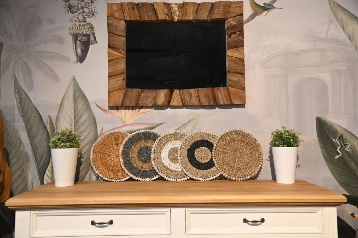 Een houten console met witte lades bevat twee potplanten en decoratieve geweven placemats in natuurlijke tinten. Erboven hangt een rustiek houten schoolbord. Het behang bevat botanische en architecturale illustraties.