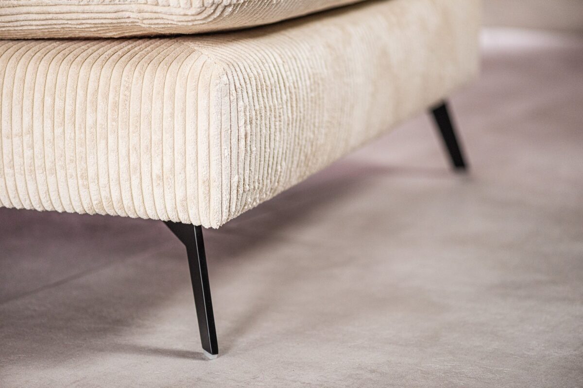 Close-up van een beige corduroy bank met zwarte metalen poten op een lichtgrijze vloer. De afbeelding richt zich op de textuur en het ontwerp van de stof en de strakke, moderne bankpoten.