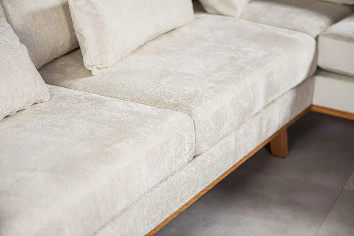 Close-up van een beige stoffen bank, met een hoekgedeelte met pluche kussens en lichte houten poten. De textuur en kleur van de stof zijn duidelijk zichtbaar, wat een zachte en uitnodigende uitstraling creëert. De vloer bestaat uit lichtgrijze tegels.