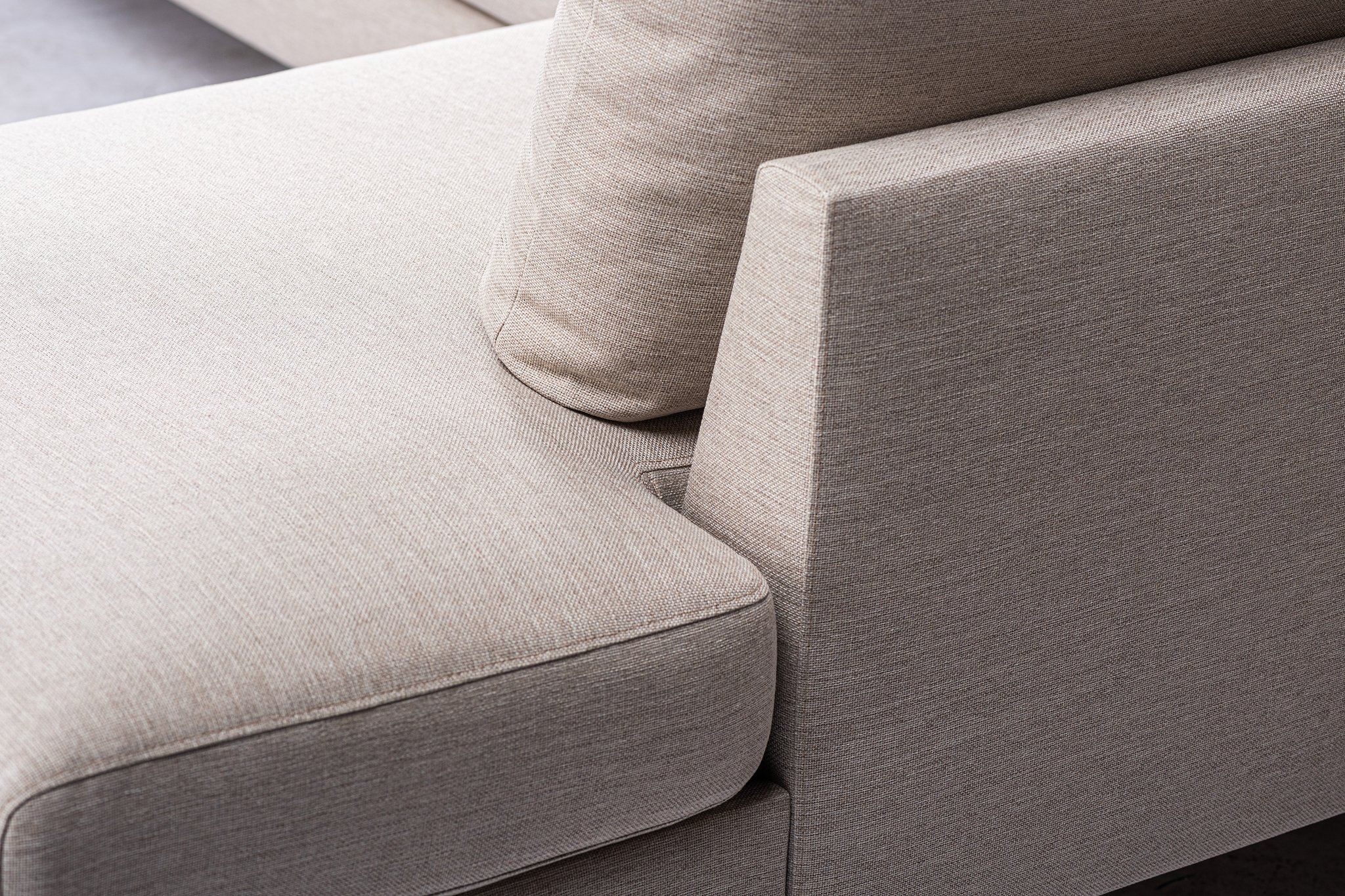 Close-up van een beige stoffen bank, met de nadruk op de strakke, scherpe lijnen waar de armleuning en het kussen elkaar ontmoeten. De textuur van de bekleding is zichtbaar, wat een minimalistisch ontwerp benadrukt.