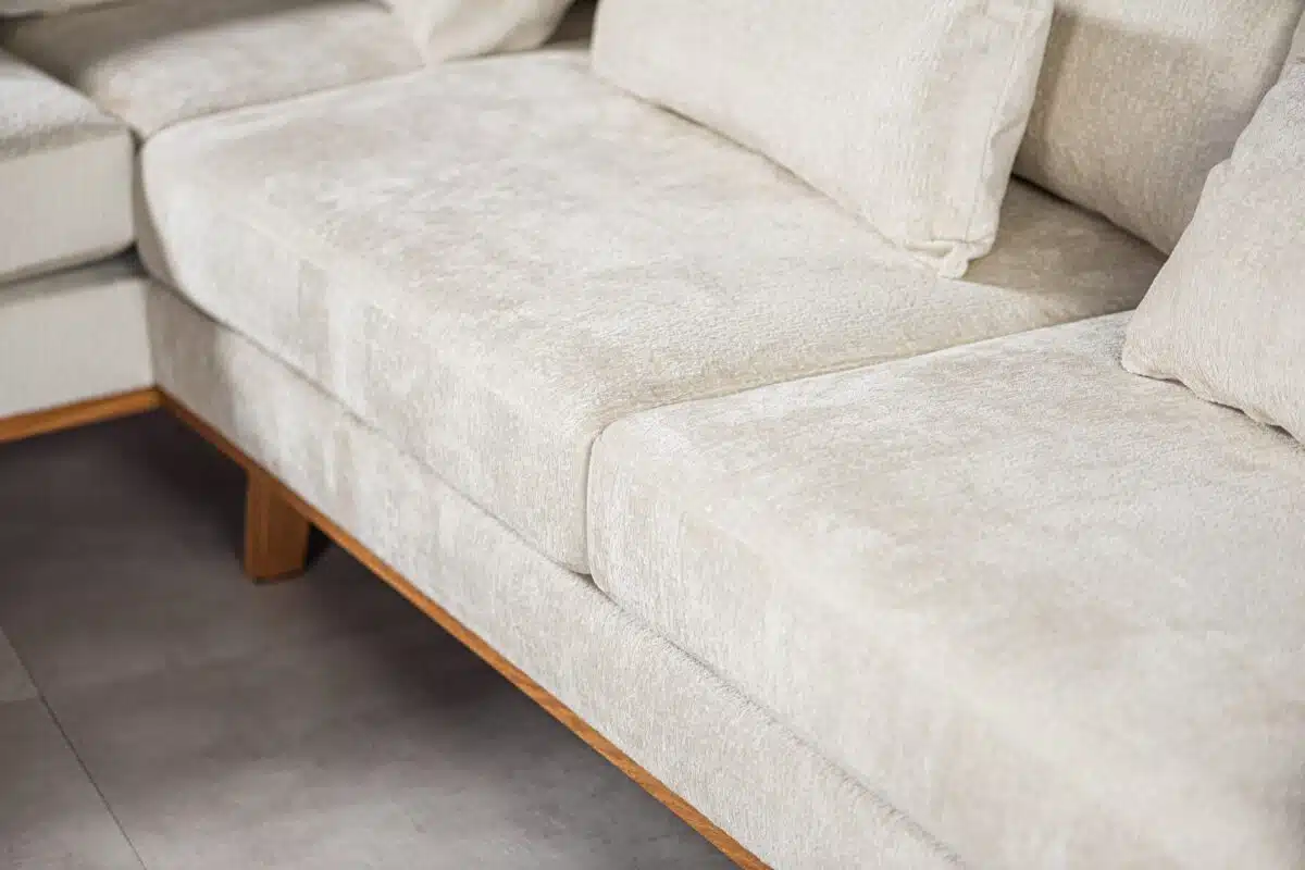 Close-up van een beige sectionele bank met structuurstof en pluche kussens. De bank heeft houten poten en staat op een lichtgekleurde tegelvloer.