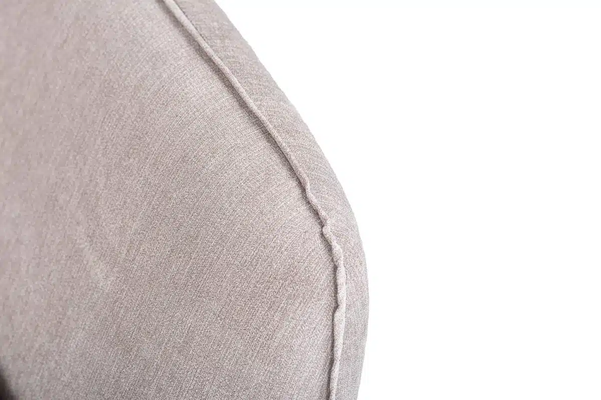 Close-up van de hoek van een beige stoffen bank met zichtbare stiksels. De textuur van de bekleding en het naaddetail zijn prominent aanwezig tegen een witte achtergrond.