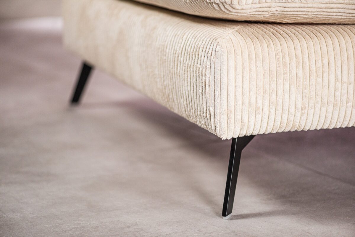 Close-up van een crèmekleurige corduroy hoekbank, met de nadruk op de textuur van de stof. De bank heeft slanke zwarte metalen poten die rusten op een gladde, lichtgekleurde vloer. Het ontwerp suggereert een moderne en minimalistische stijl.