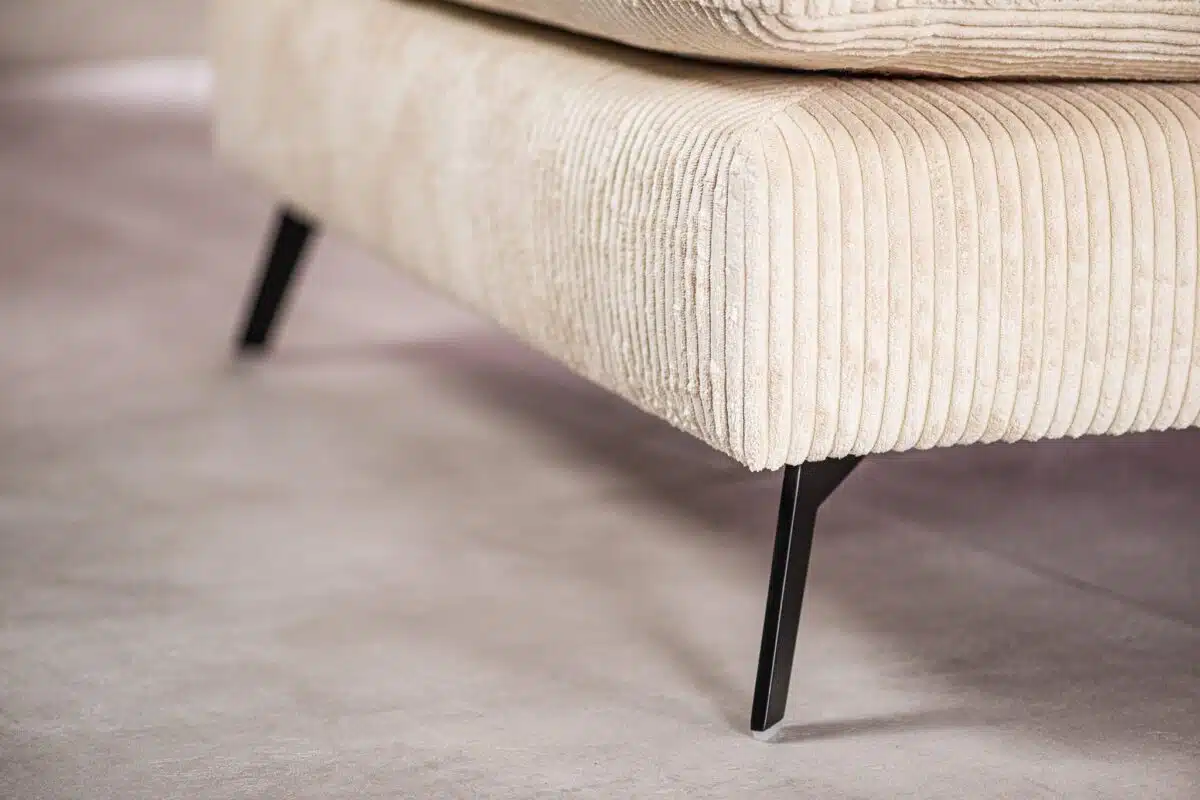 Close-up van een crèmekleurige corduroy hoekbank, met de nadruk op de textuur van de stof. De bank heeft slanke zwarte metalen poten die rusten op een gladde, lichtgekleurde vloer. Het ontwerp suggereert een moderne en minimalistische stijl.