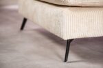 Close-up van een crèmekleurige corduroy hoekbank, met de nadruk op de textuur van de stof. De bank heeft slanke zwarte metalen poten die rusten op een gladde, lichtgekleurde vloer. Het ontwerp suggereert een moderne en minimalistische stijl.
